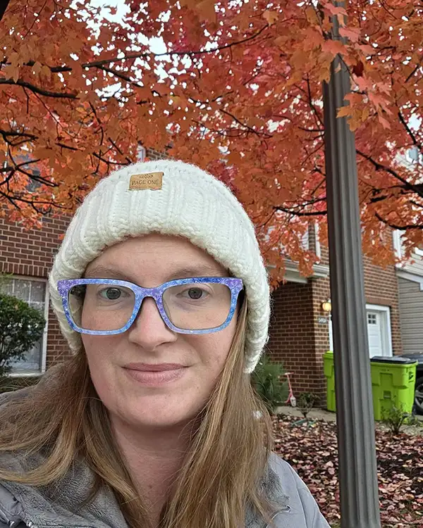 A woman in a white knit beanie and patterned glasses smiles in front of a tree with red leaves.