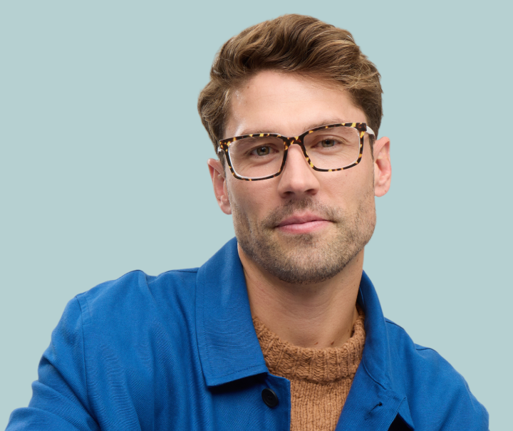 A man with light brown hair wearing tortoiseshell glasses, a brown sweater, and a blue jacket.