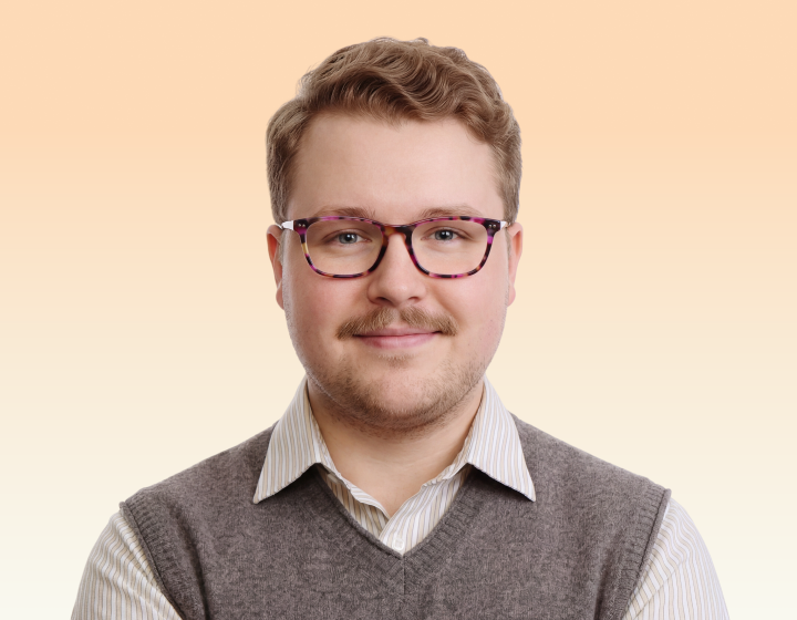 A man with wavy hair, a mustache, and glasses, wearing a sweater vest and collared shirt.