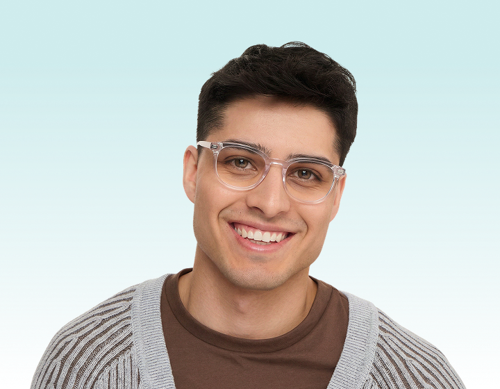A smiling man with dark hair wears clear-framed glasses, a brown shirt, and a grey striped cardigan.