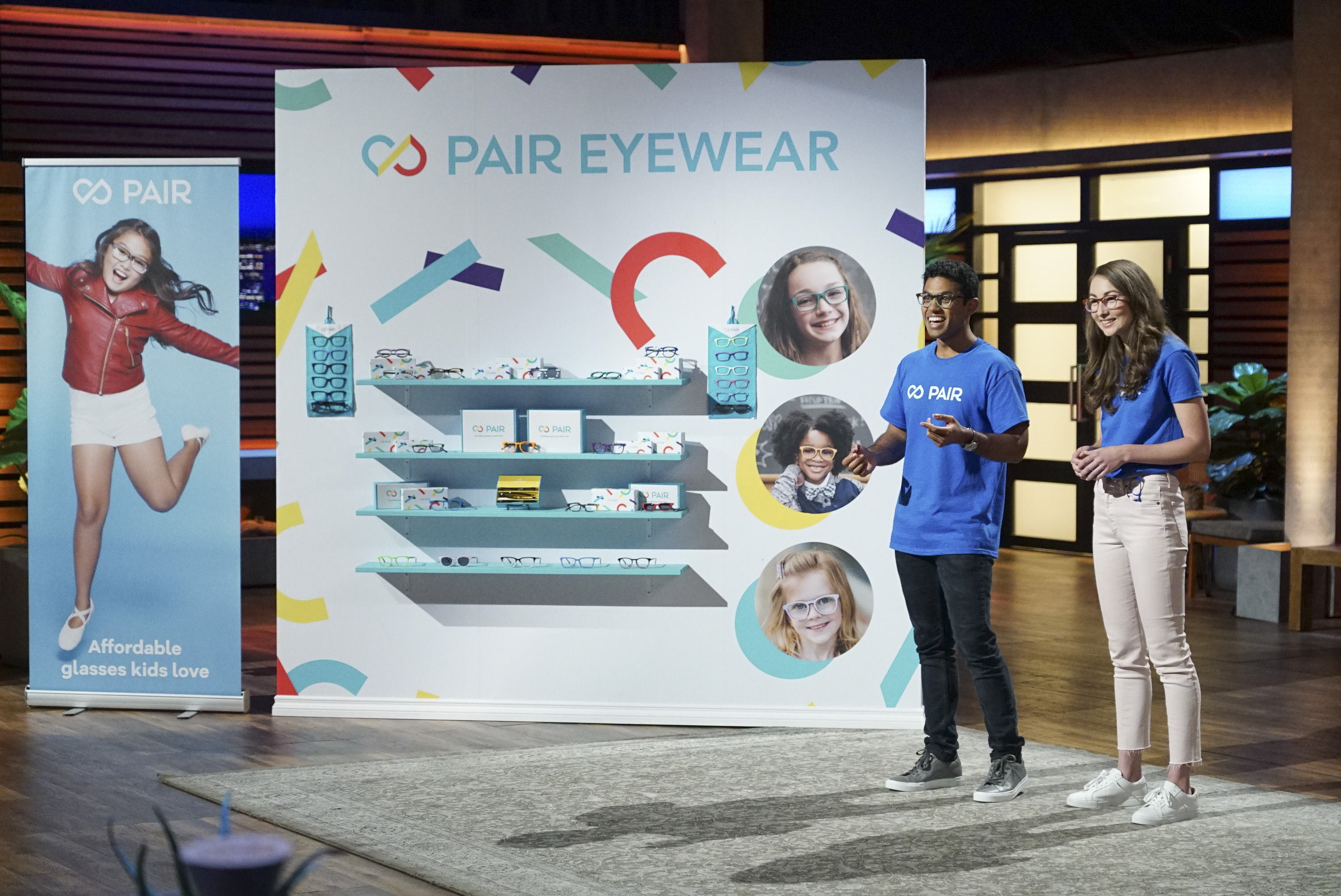 A man and a woman present Pair Eyewear on a stage with a display of glasses.