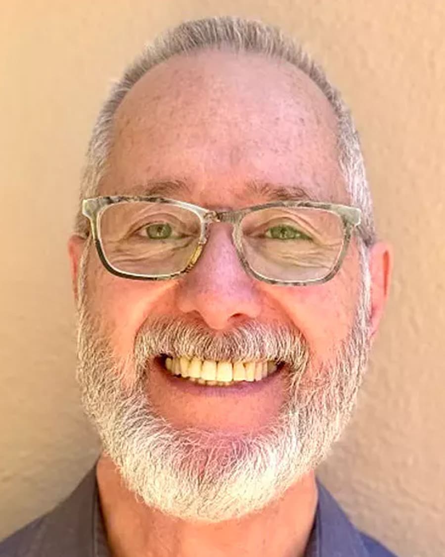 Smiling man with glasses and a beard against a beige background.