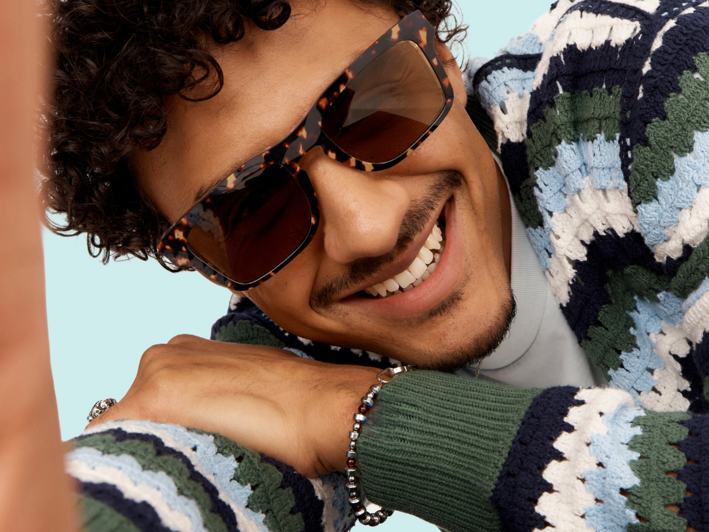 Smiling person wearing sunglasses and a colorful sweater, leaning on their arm.