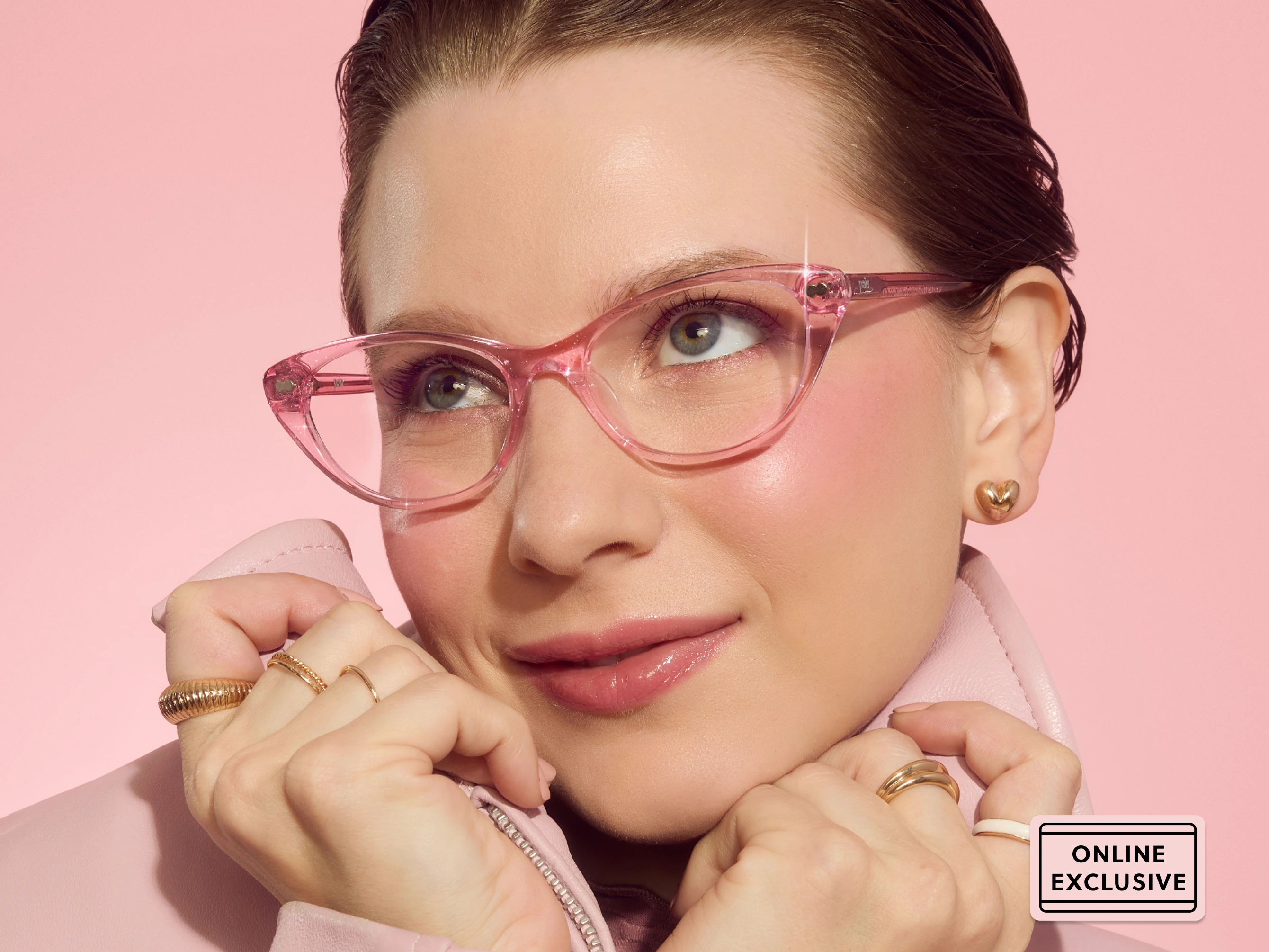 Person wearing pink glasses and coat with gold earrings and rings, pink background.
