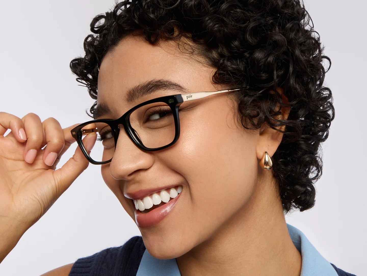 A smiling woman with short curly hair adjusts her black and gold eyeglasses.