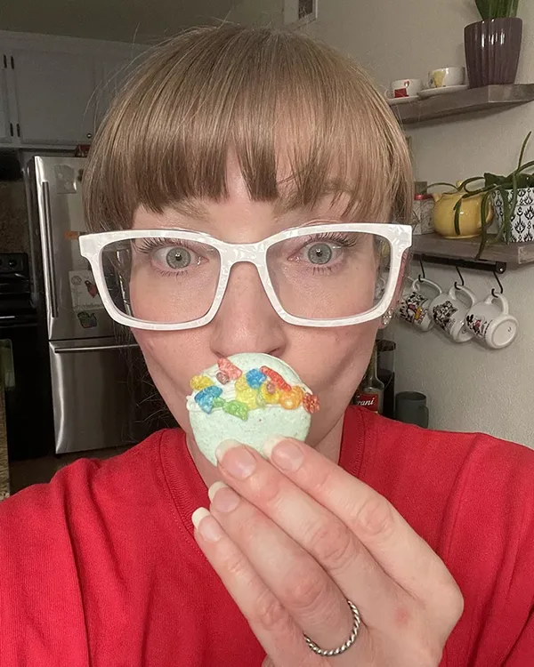 A woman in white glasses holds a light green macaron with colorful gummy worms up to her mouth.