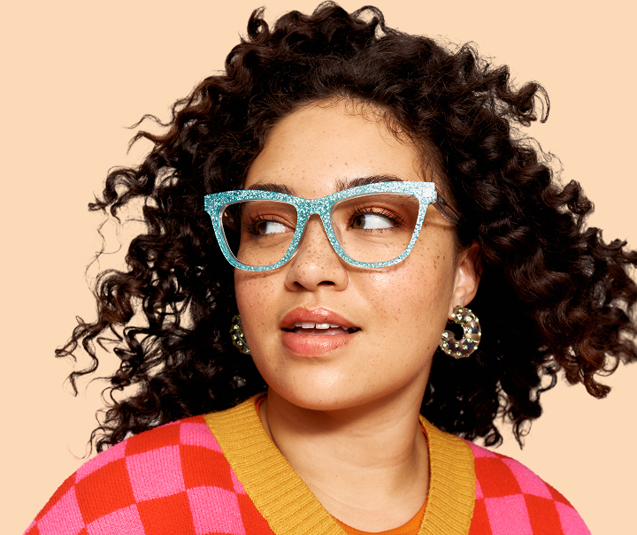 A woman with curly hair and freckles wearing sparkly blue glasses and a colorful checkered sweater.