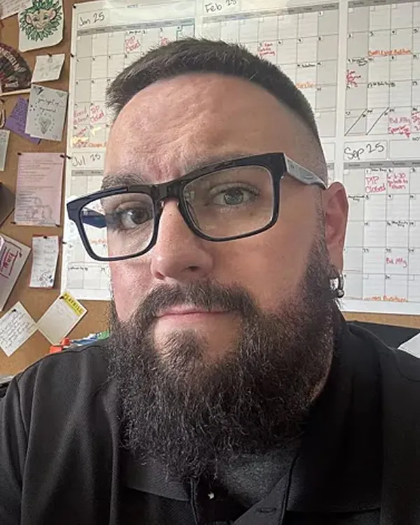 A close-up photo of a man with a dark beard and black-rimmed glasses, in front of a wall calendar.