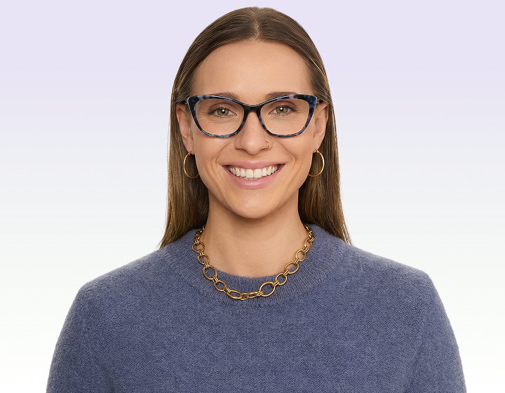 A smiling woman with brown hair wears blue patterned glasses, a blue sweater, and a gold chain necklace.