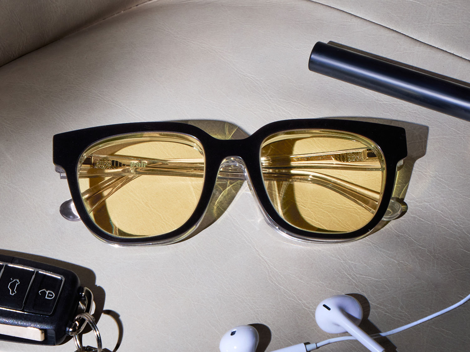 A pair of sunglasses with yellow lenses lies on a surface with a car key and earbuds nearby.