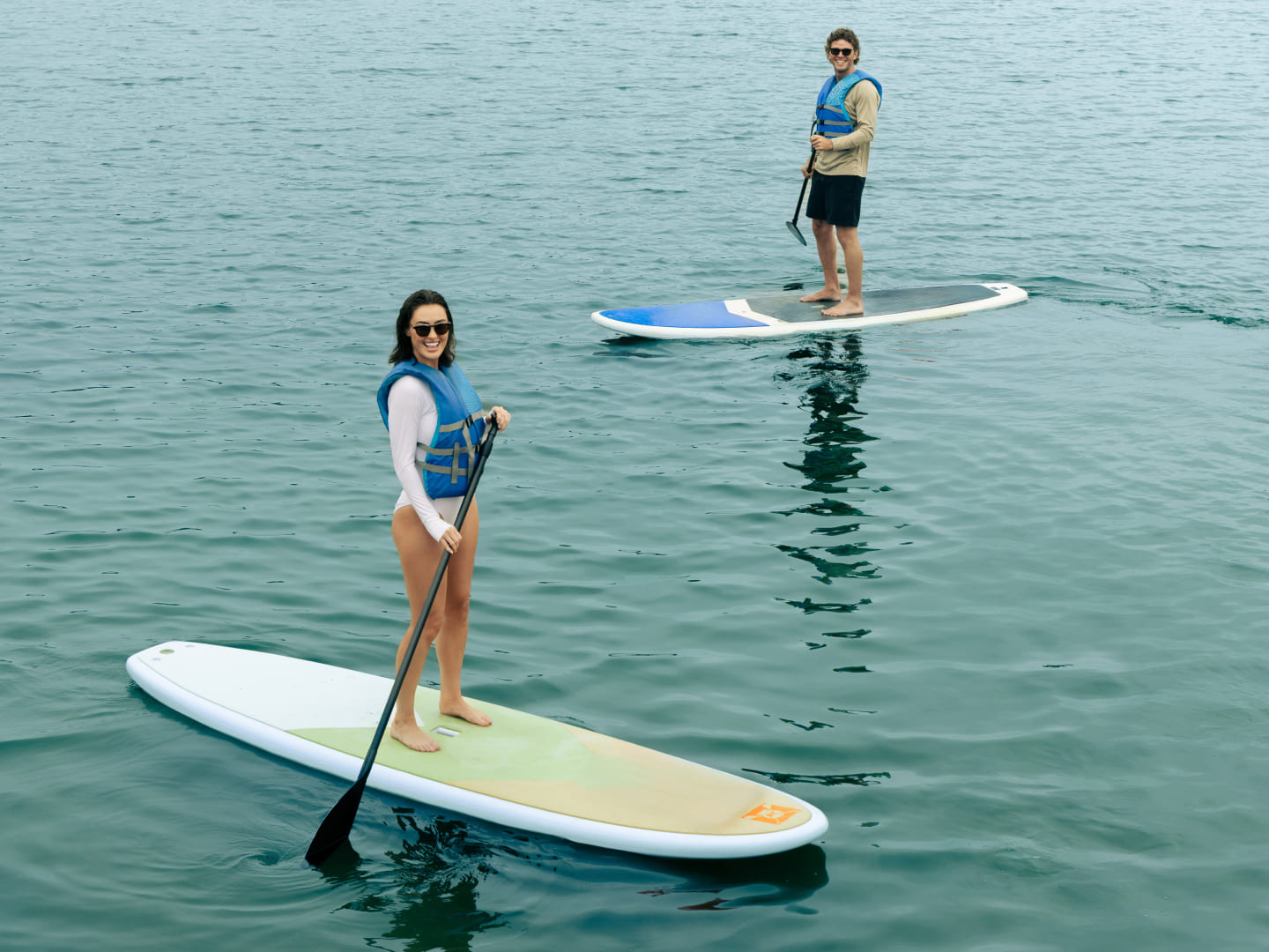 Two people paddleboarding on calm water, wearing life vests and holding paddles.