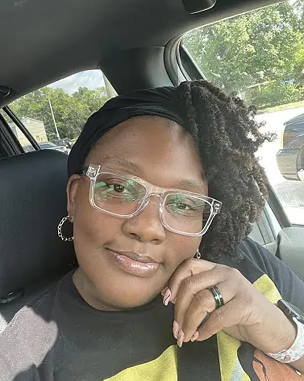 A smiling woman with glasses and a black headwrap sits in a car.