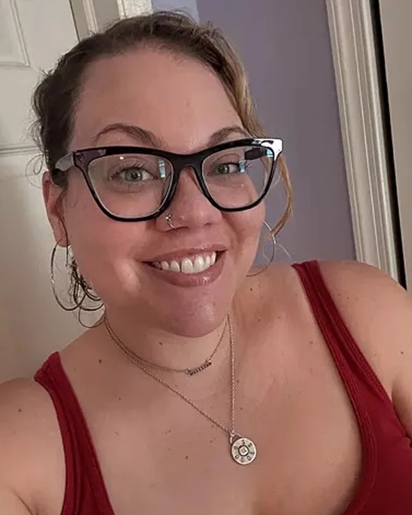 A smiling woman with light brown hair wearing black glasses, a nose ring, and a red tank top.