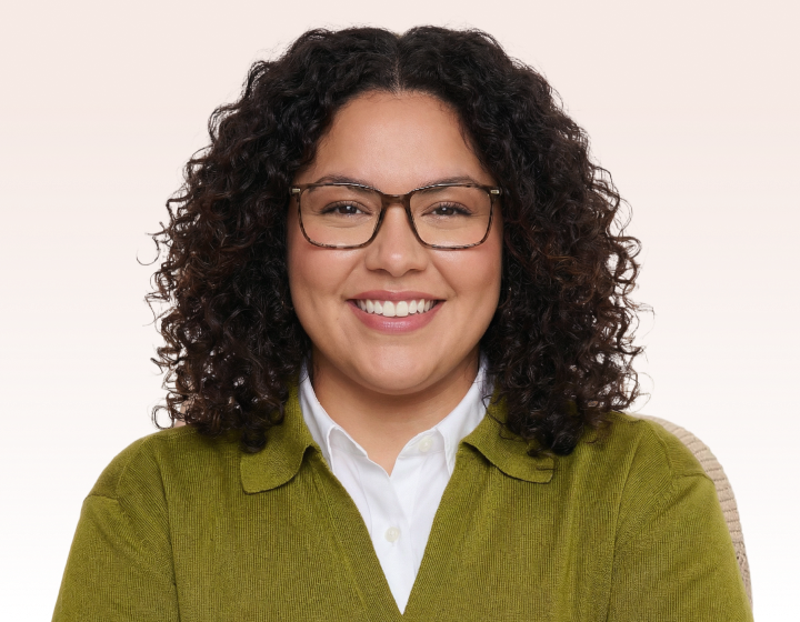 A smiling woman with dark curly hair and glasses, wearing a green cardigan over a white shirt.