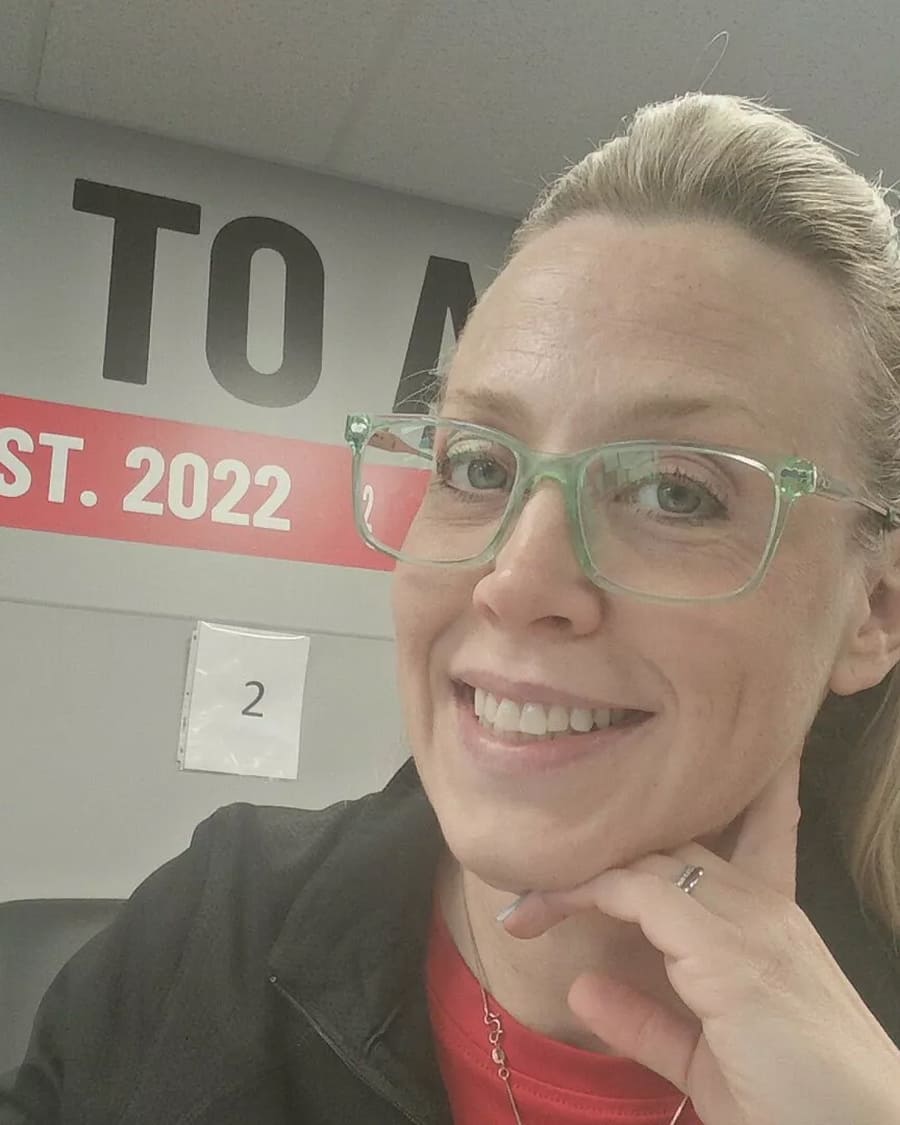 Person with glasses smiling, sitting indoors with a sign reading 'EST. 2022' in the background.