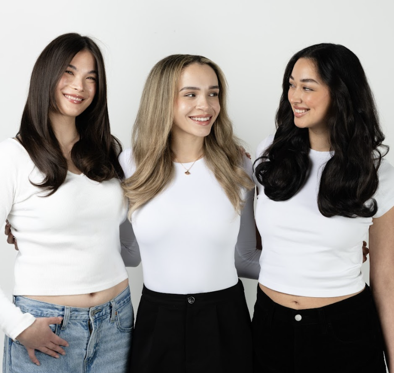 Three smiling women with different hair colors and white tops stand together with their arms around each other.