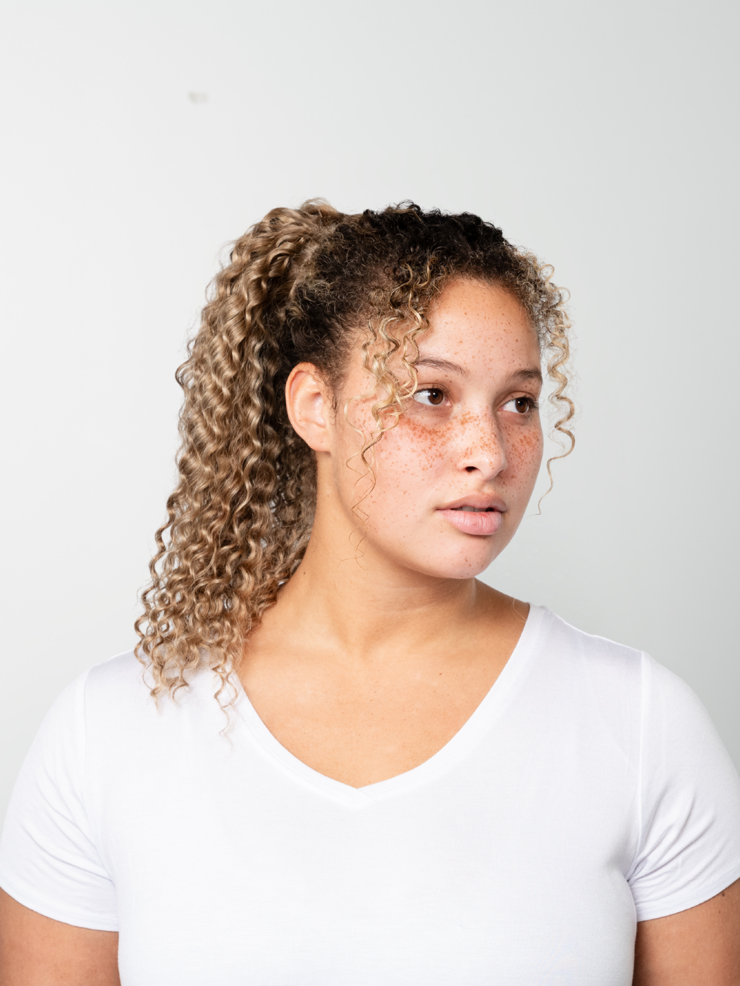 Model with curly light brown hair styled in a high voluminous ponytail, wearing a white t-shirt.