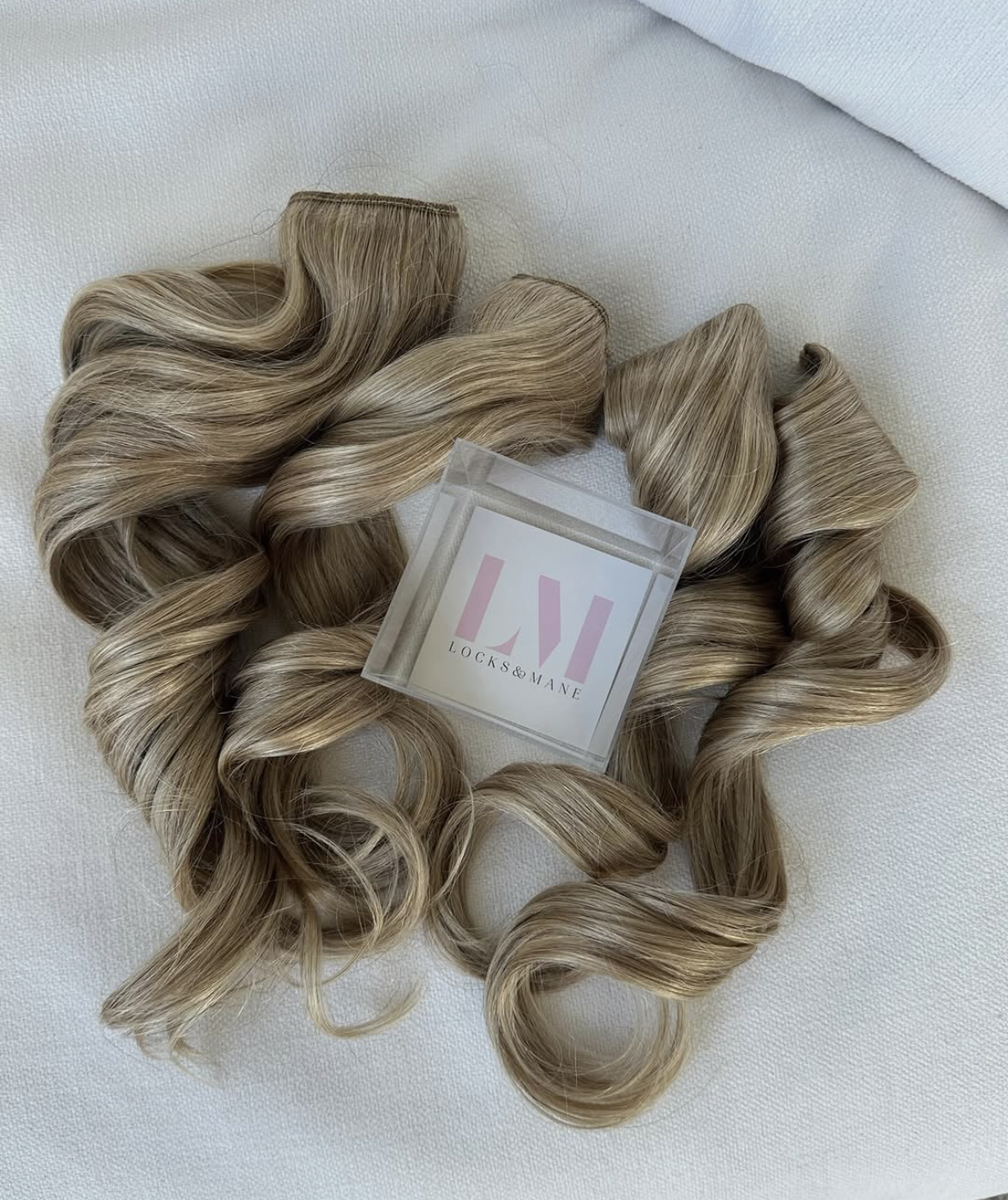 Wavy, highlighted blonde hair extensions from Locks & Mane displayed on a white textured surface.