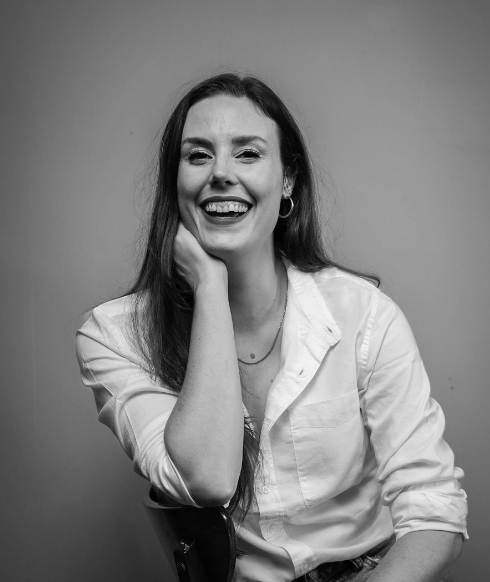 Black and white photo of a smiling person with long hair, wearing a white shirt, sitting on a chair.