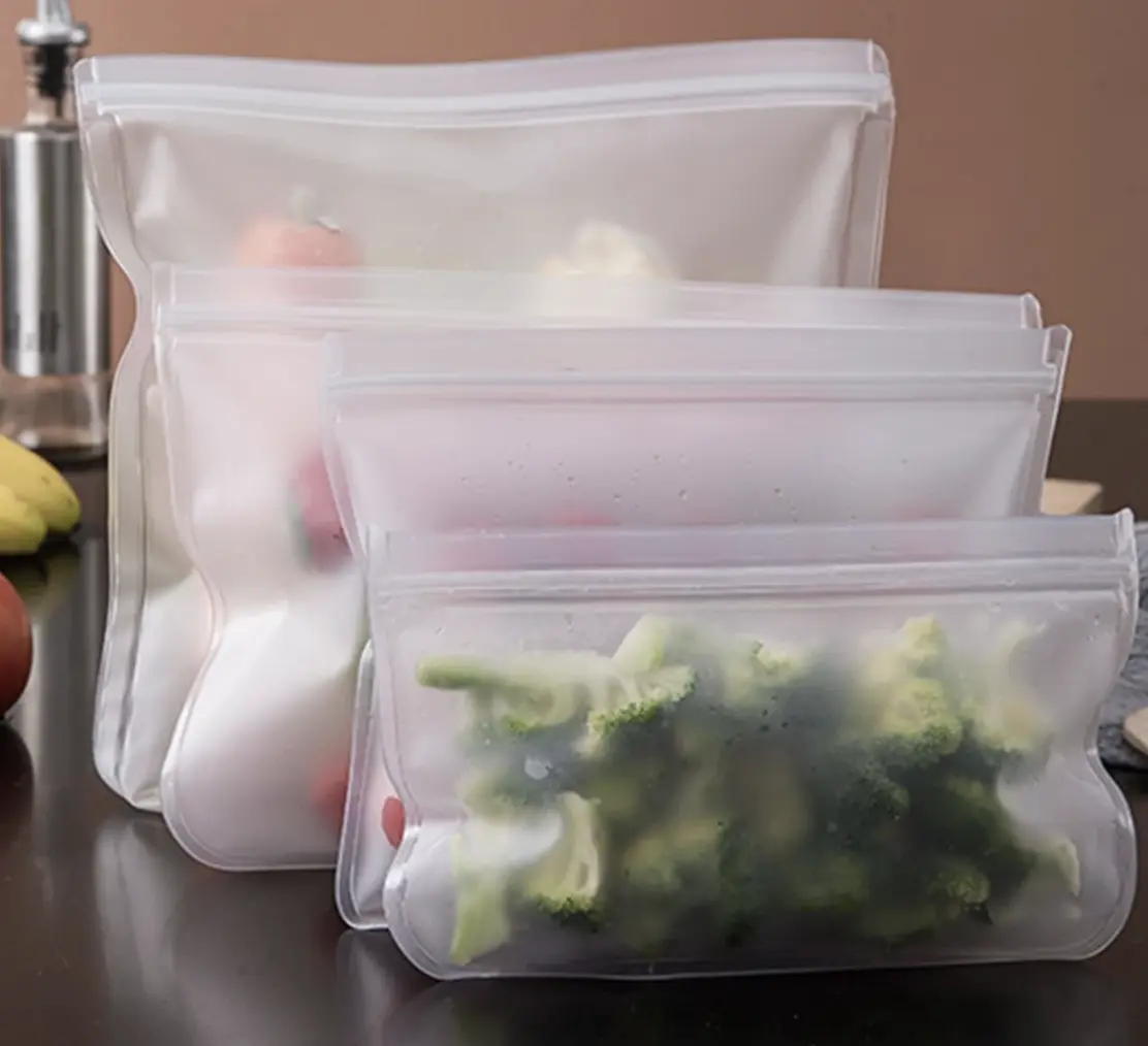Three translucent, reusable zippered bags of different sizes filled with vegetables like broccoli on a countertop.