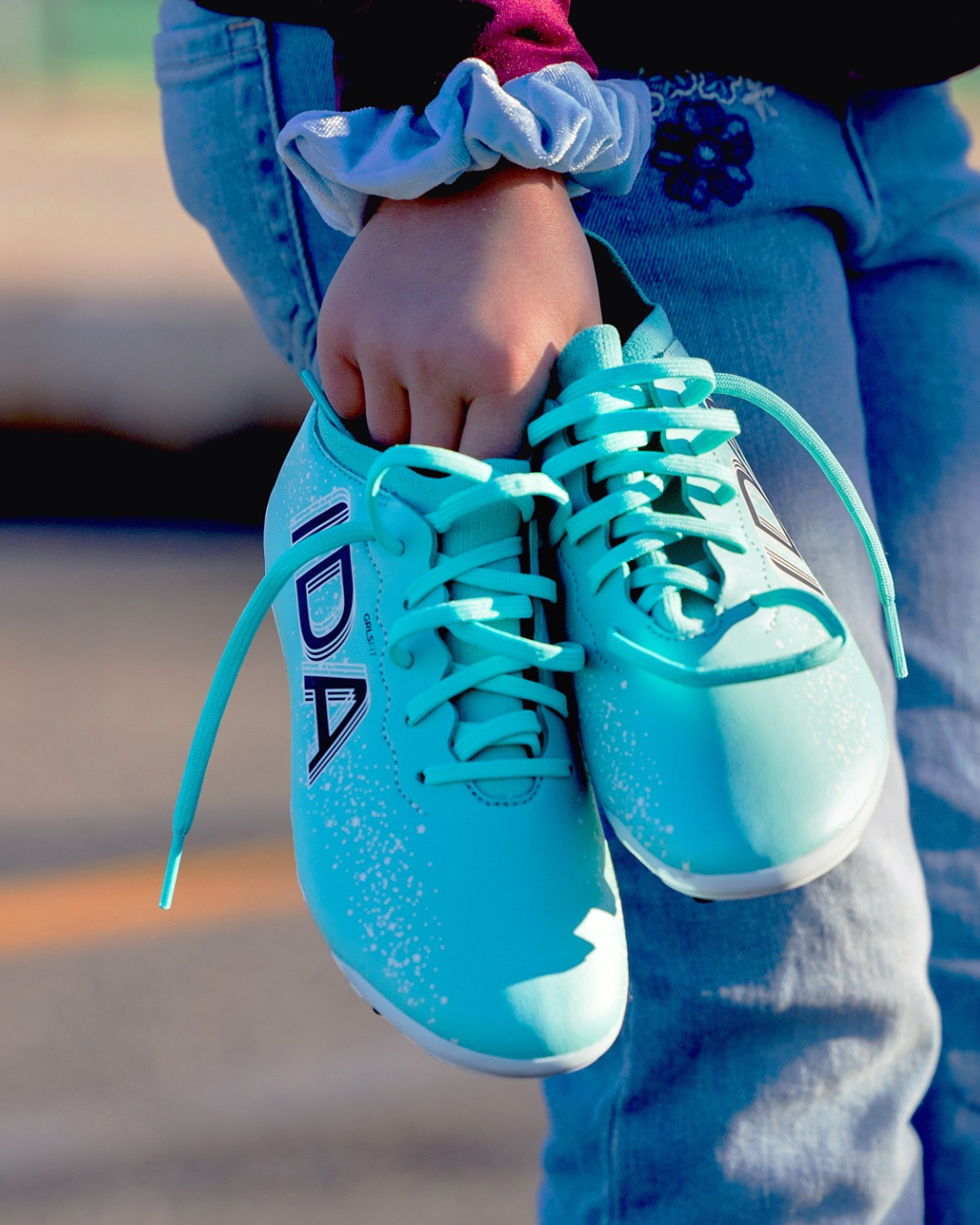 Child holding turquoise sports shoes with laces, wearing jeans and a scrunchie.