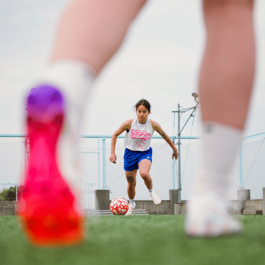 A woman dribbles a soccer ball on a green field, viewed from a low angle behind another player.