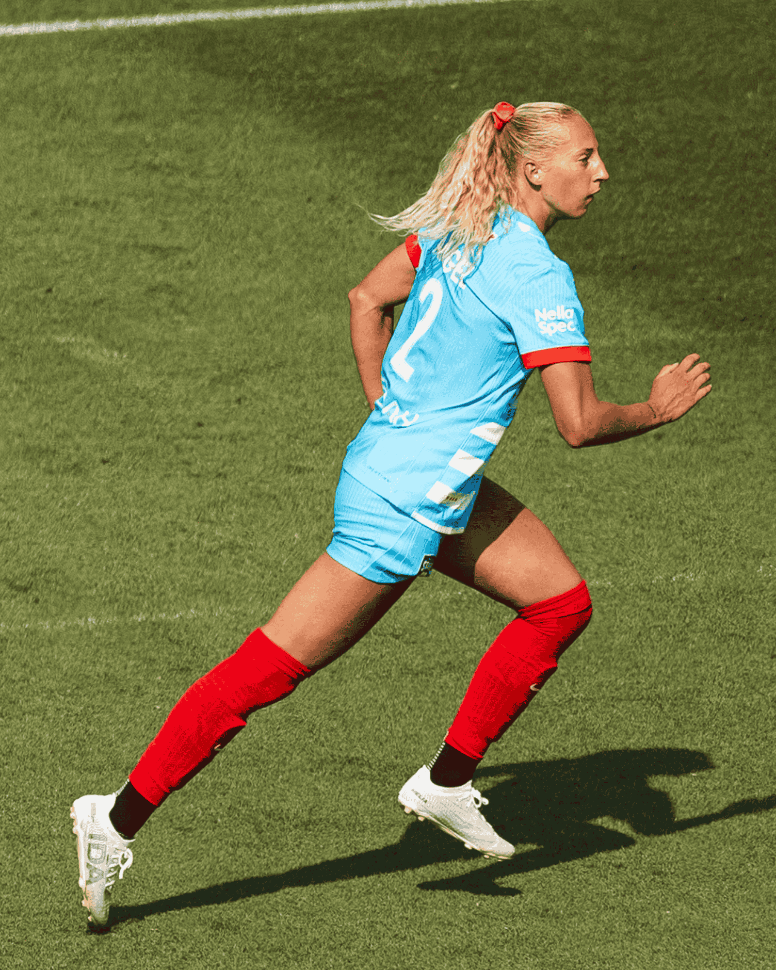 A female soccer player in a light blue uniform and red socks runs across a green field.