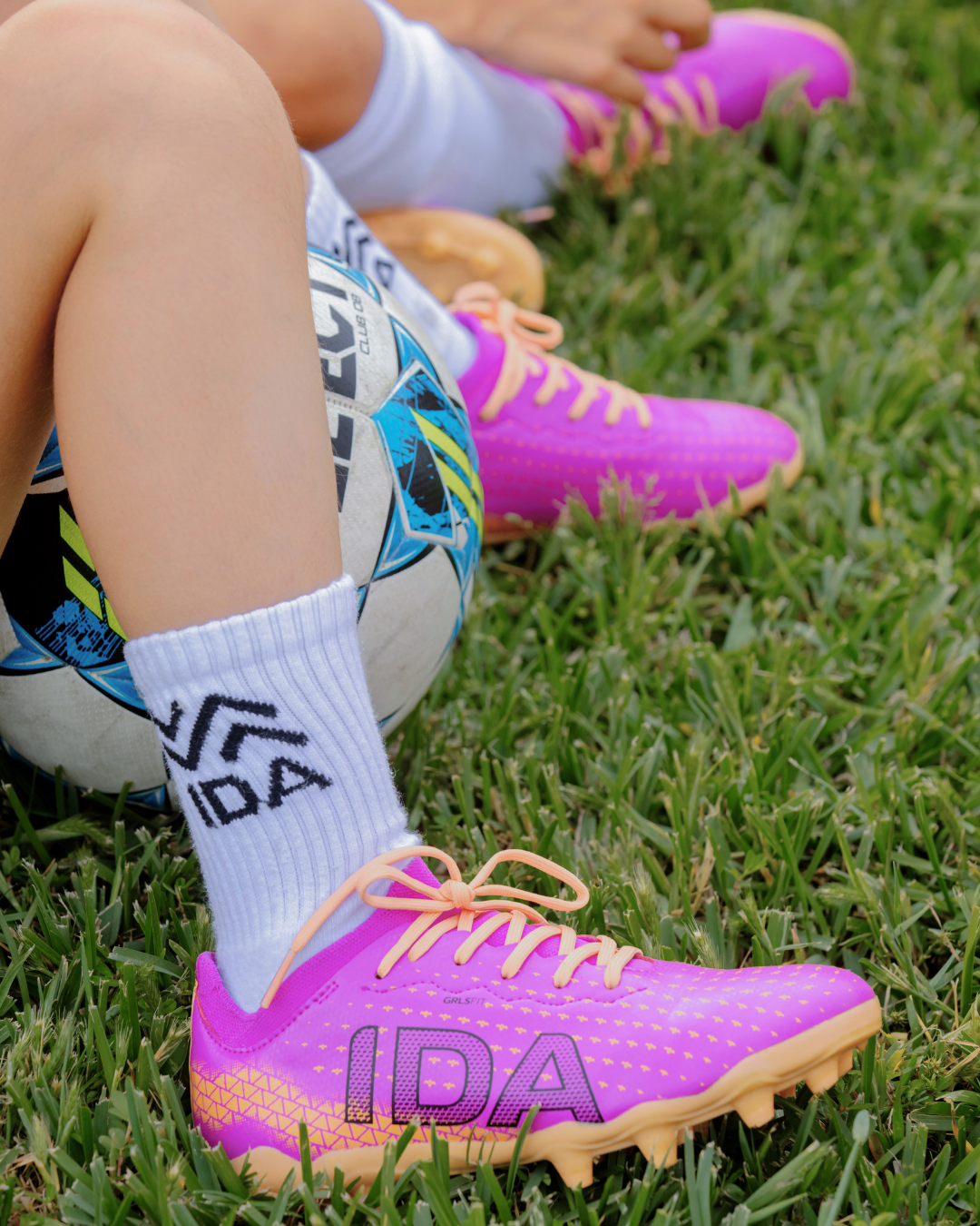 A close-up of a person wearing pink soccer cleats and white socks, sitting in grass next to a soccer ball.