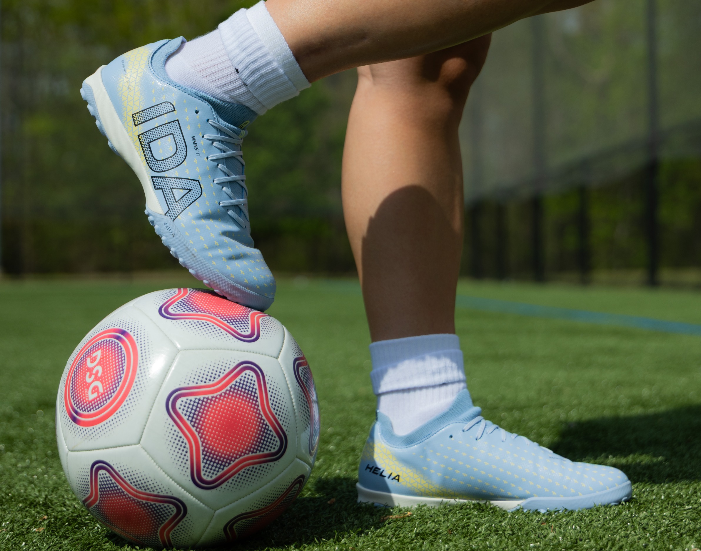 A close-up of a person's legs wearing light blue soccer cleats and white socks on artificial turf.