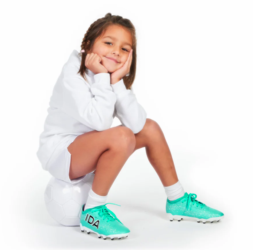 Child sitting on a soccer ball, wearing turquoise cleats and a white outfit.