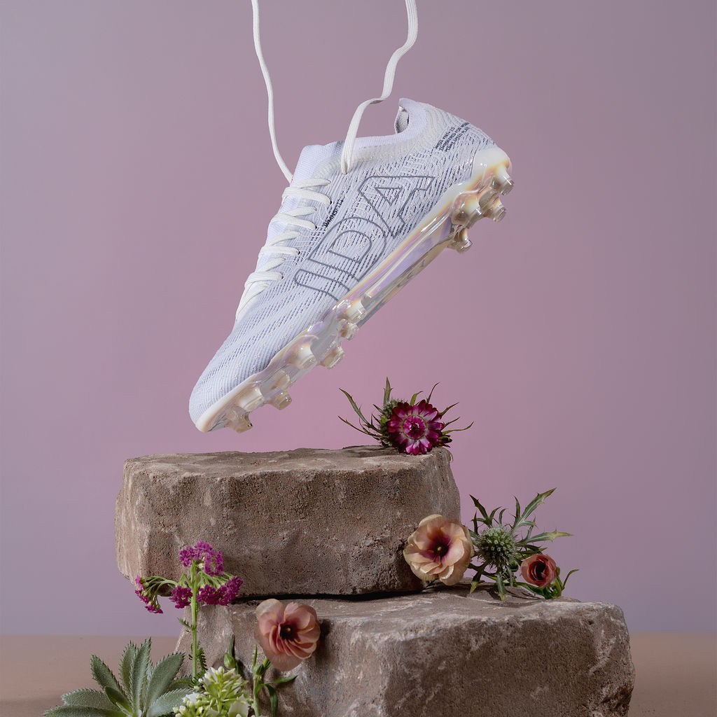 White soccer cleat suspended above stone blocks with flowers, against a pink background.