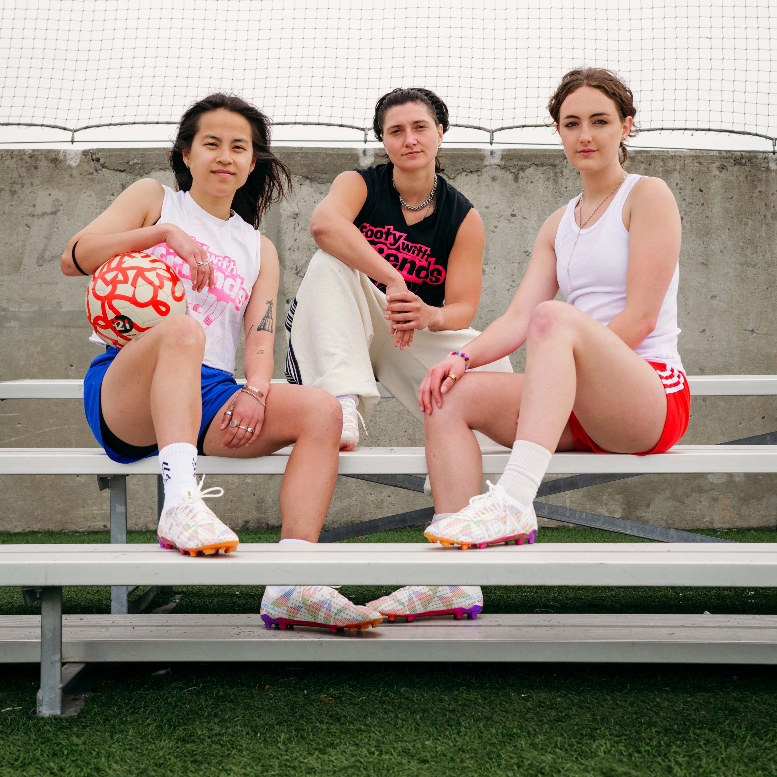 Three women in athletic wear sit together on a bleacher bench, one holding a soccer ball.