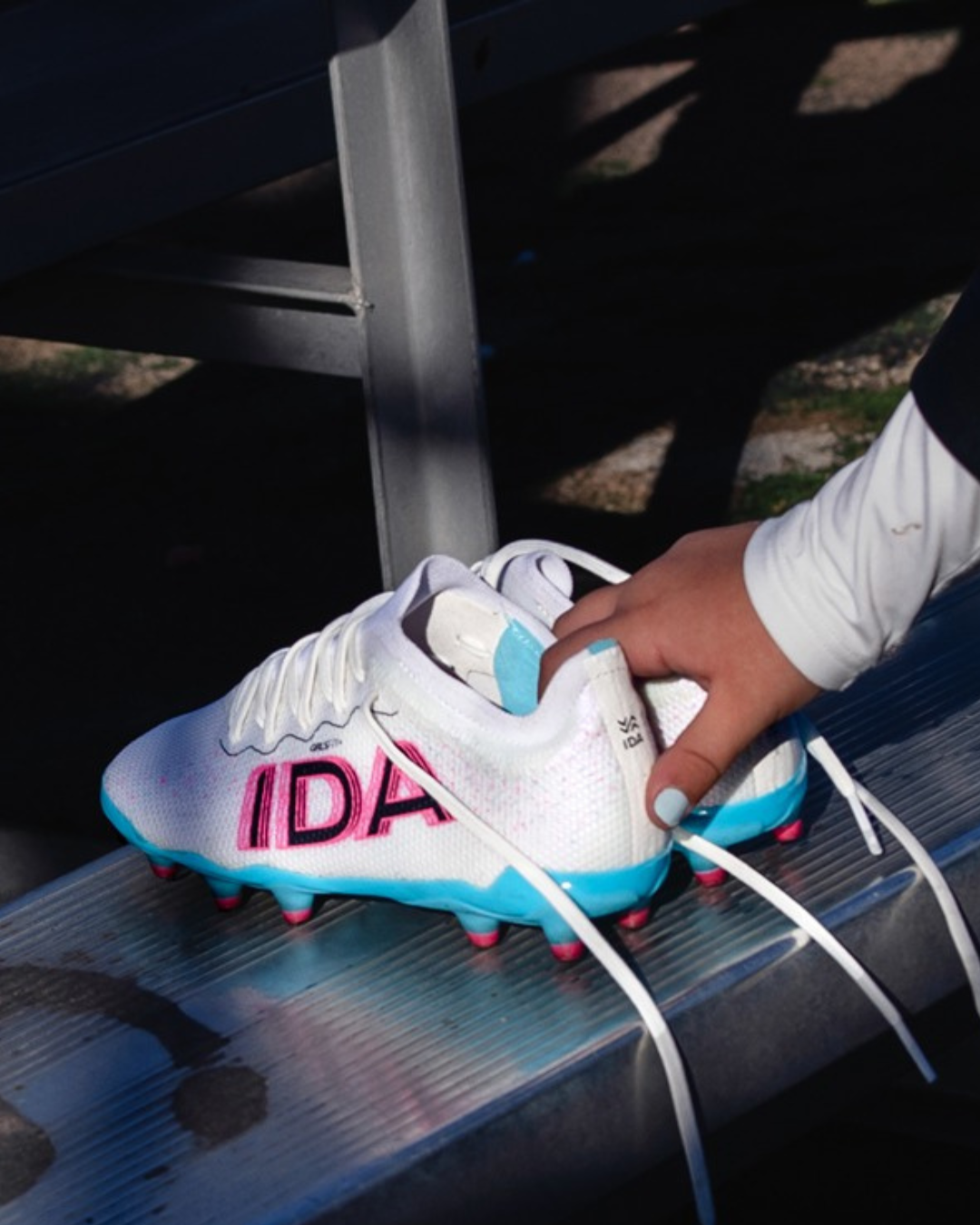 Person holding white soccer cleats with blue soles and 'IDA' text on a bench.