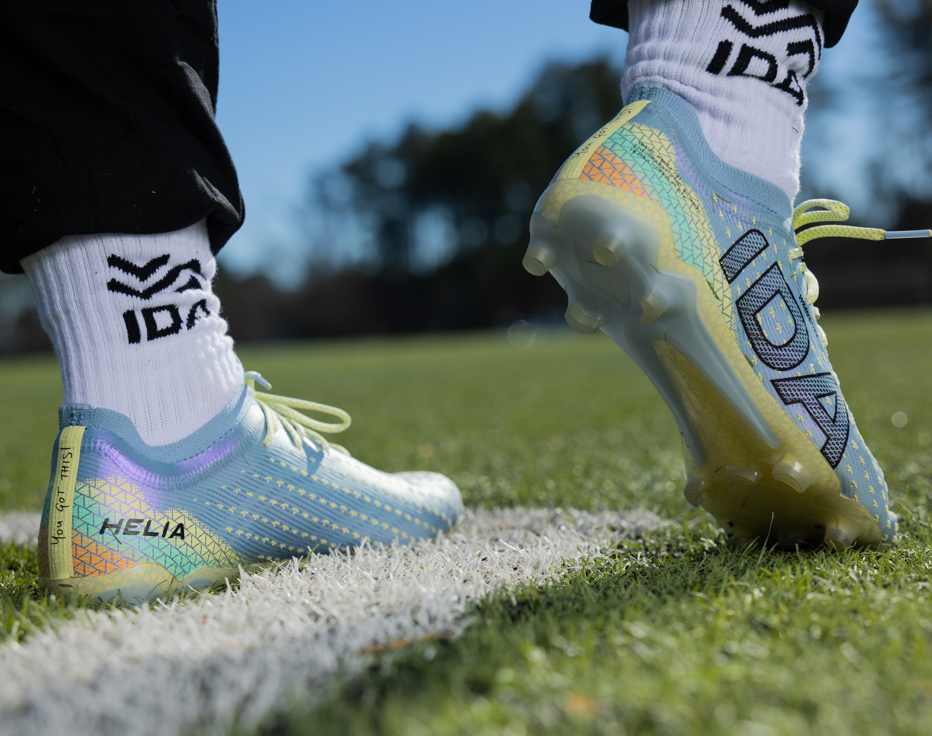 A close-up, low-angle view of a light blue IDA soccer cleat on a foot in a white sock.