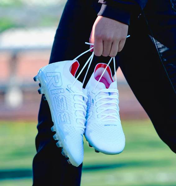 Person holding white soccer cleats with 'RISE' text, outdoors.
