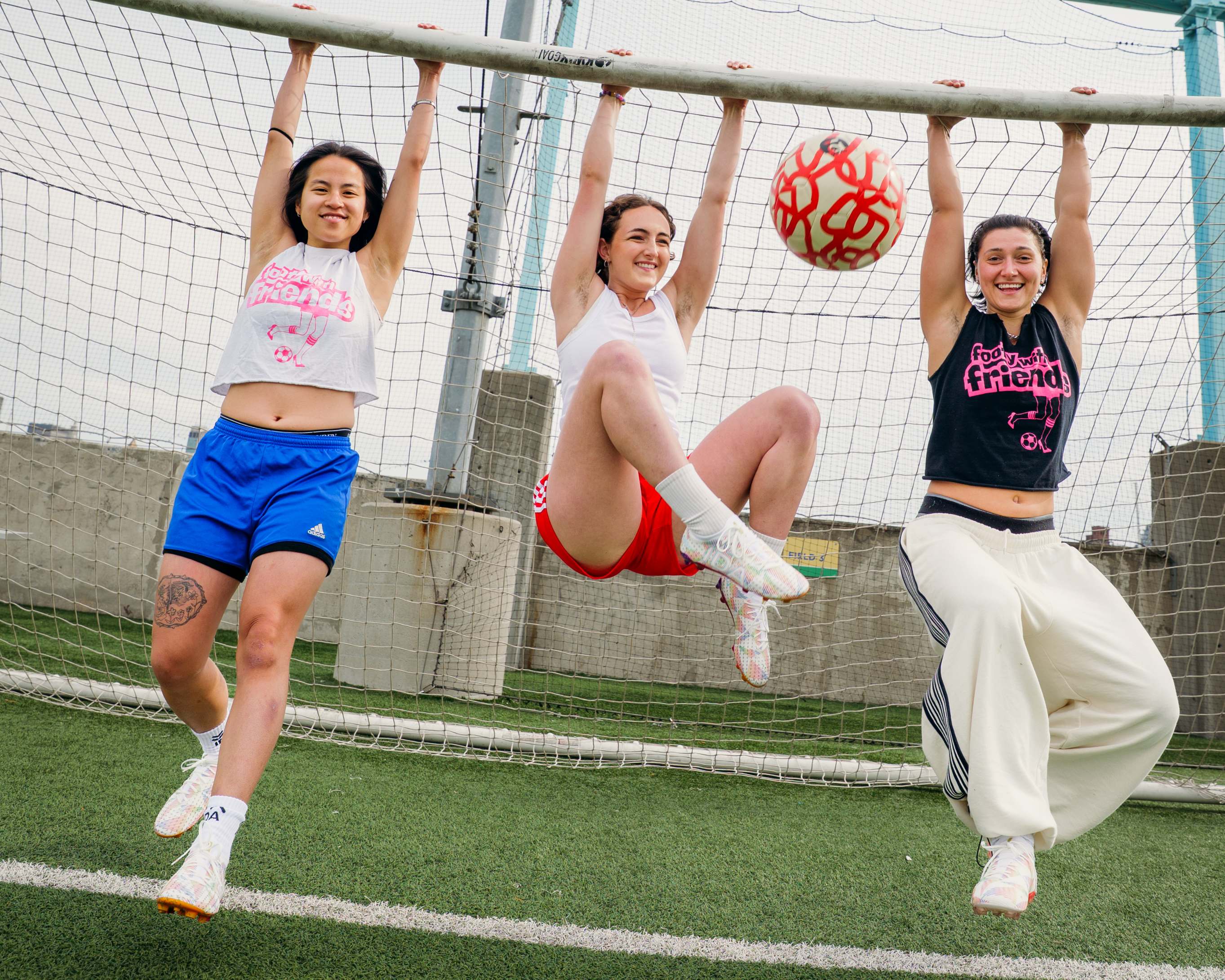 Three smiling women playfully hang from the crossbar of a soccer goal on a turf field.