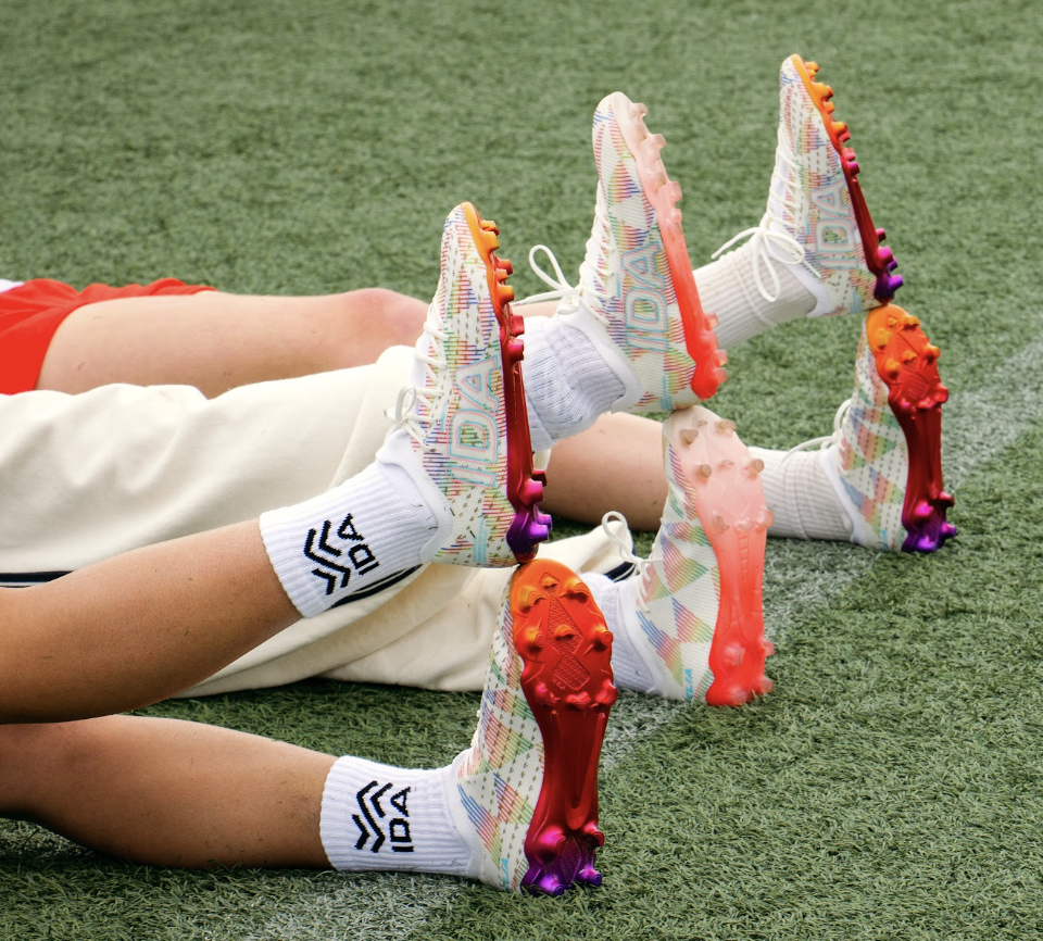 Three people lying on a grass field, showing off their colorful soccer cleats and white socks.