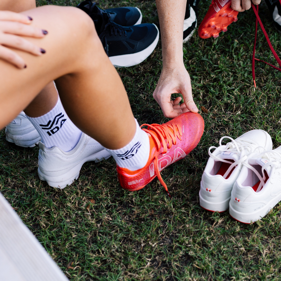 Person tying shoelaces on bright pink sports shoes on grass, with other shoes nearby.