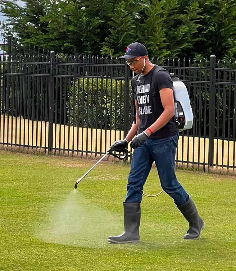 Person spraying lawn with backpack sprayer, wearing boots and gloves.
