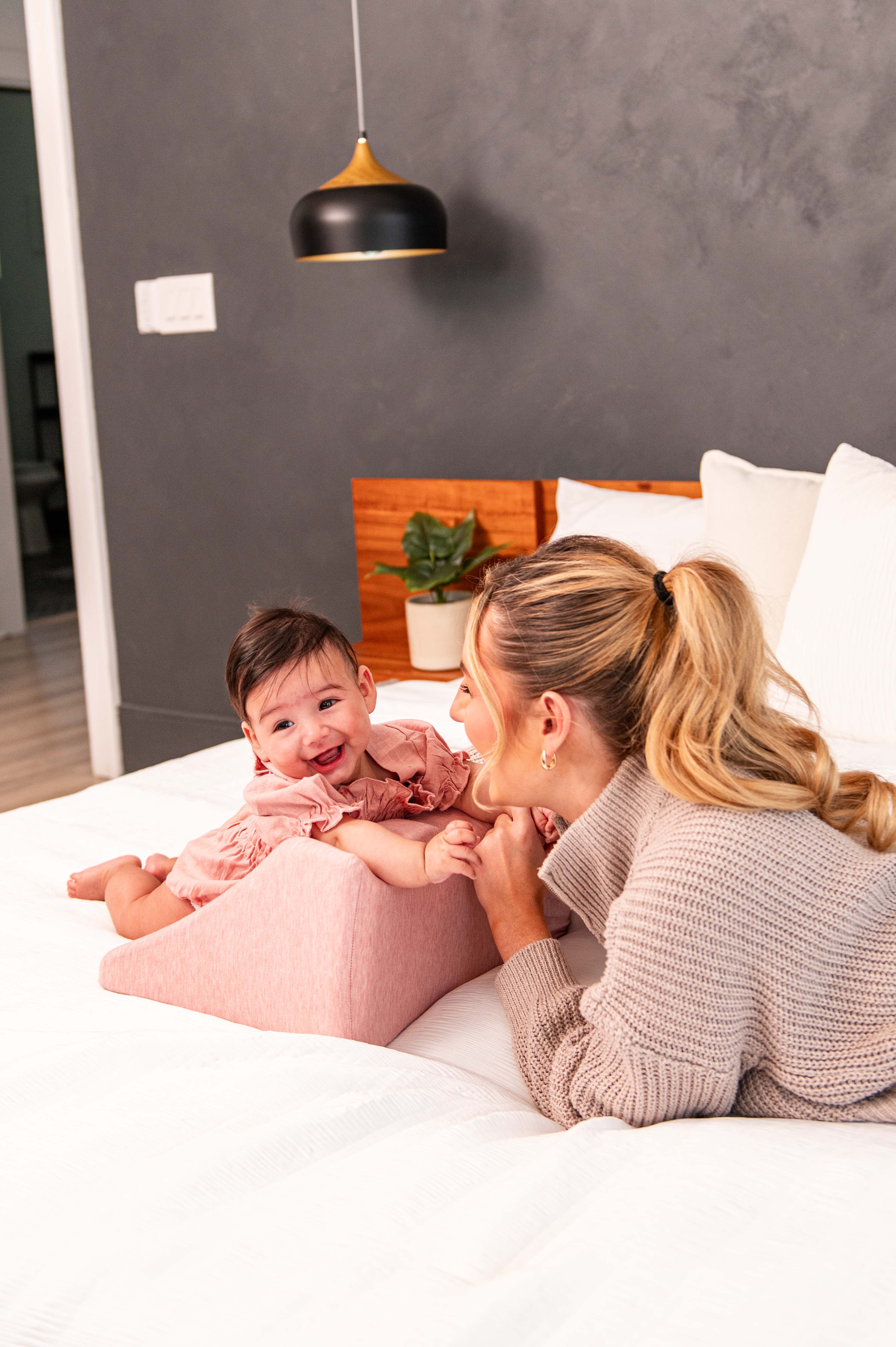 Woman and baby smiling on a bed with a pink cushion.