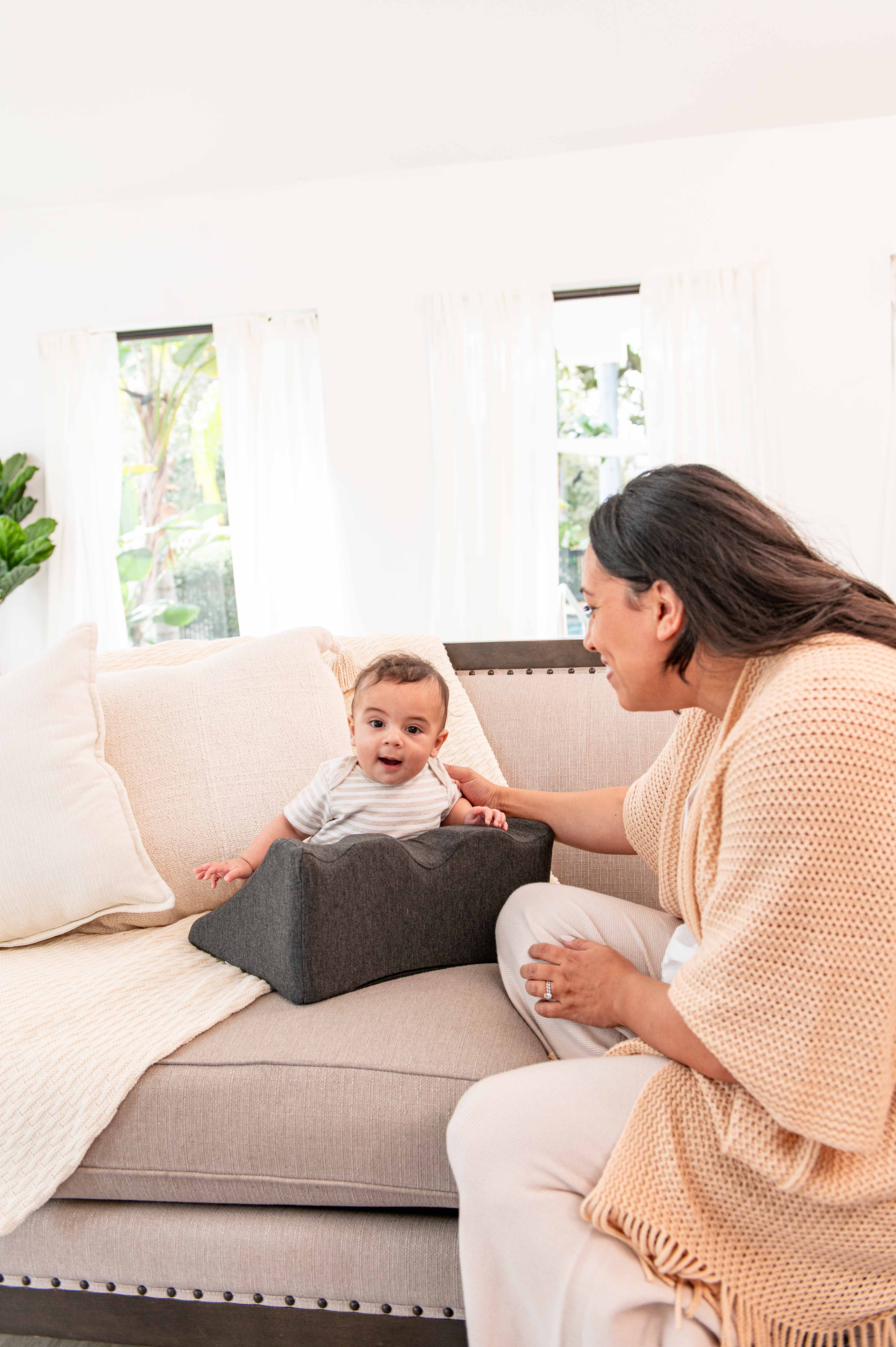 A baby sitting on a couch with an adult nearby, smiling.