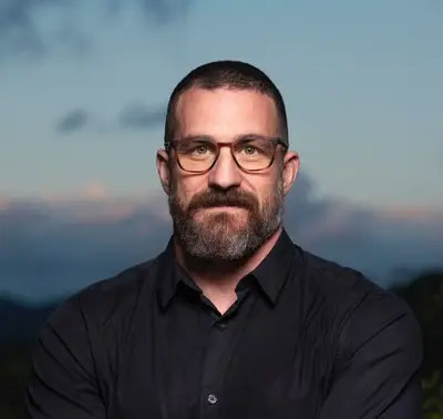A person with glasses and a beard, wearing a black shirt, stands outdoors.
