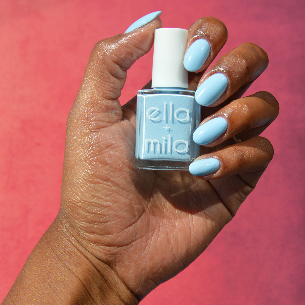 A hand with a light blue manicure holds a matching bottle of Ella + Mila nail polish.