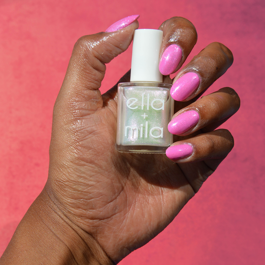 A hand with sparkly pink nails holds a bottle of iridescent Ella + Mila nail polish.