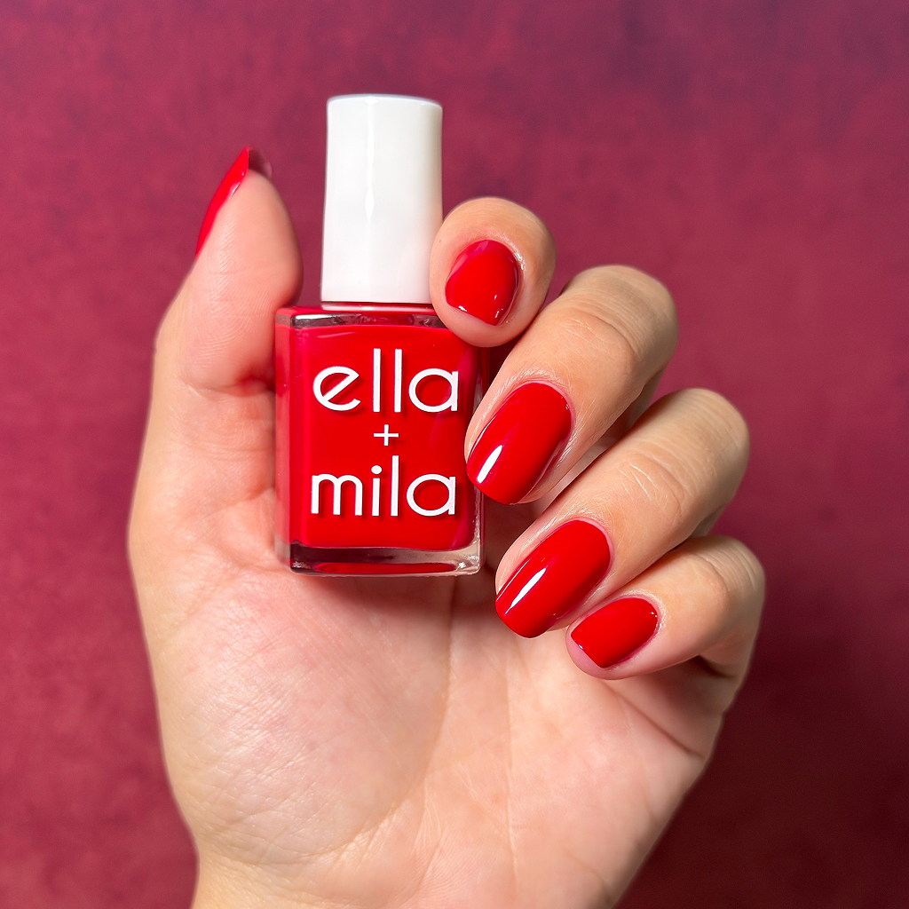 A hand with a glossy red manicure holding a bottle of Ella + Mila red nail polish.