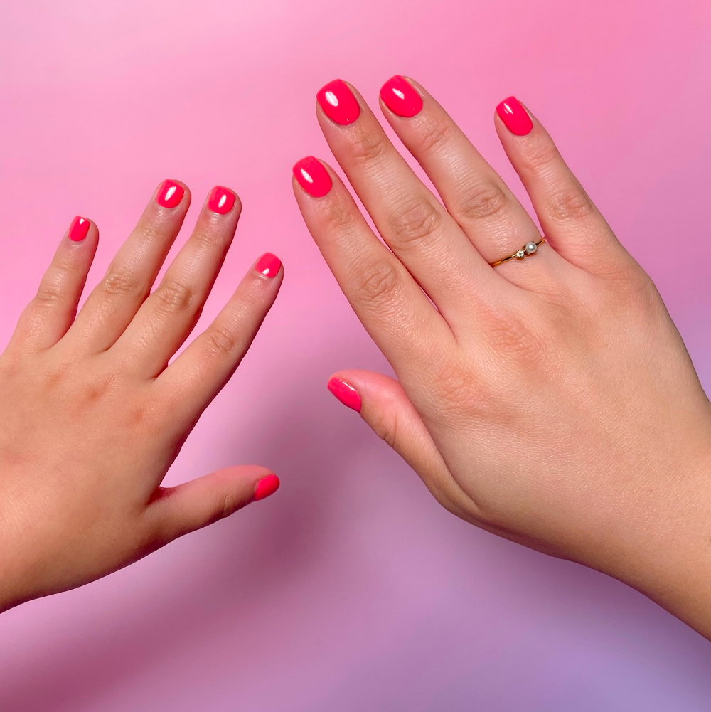 An adult's and a child's hands with matching bright pink nail polish against a pink gradient background.