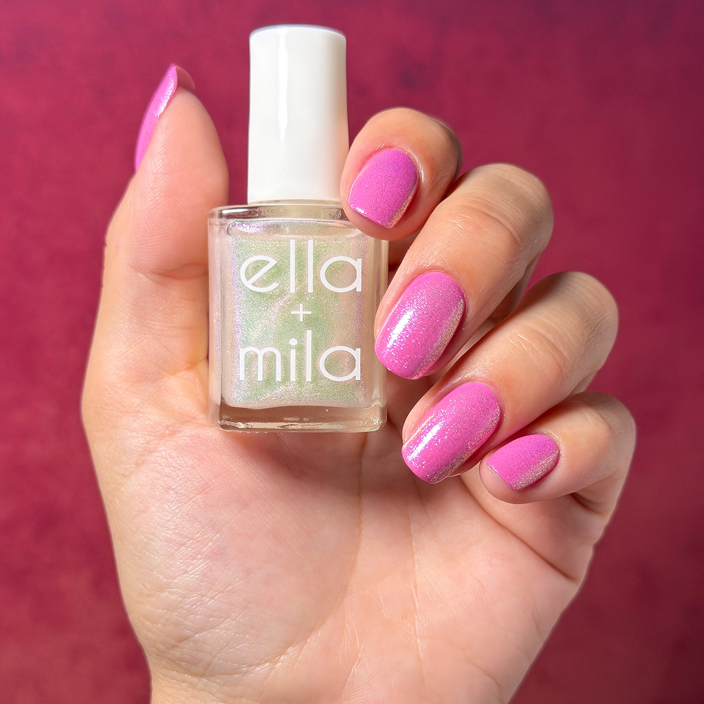 A hand with shimmery pink nails holds a bottle of Ella + Mila nail polish against a magenta background.