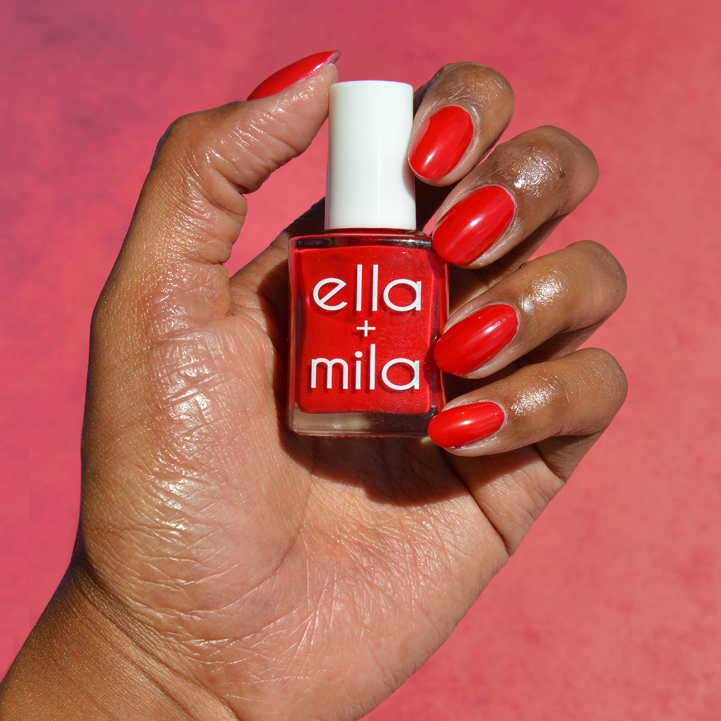 A hand with glossy red painted nails holds a bottle of ella + mila red nail polish.