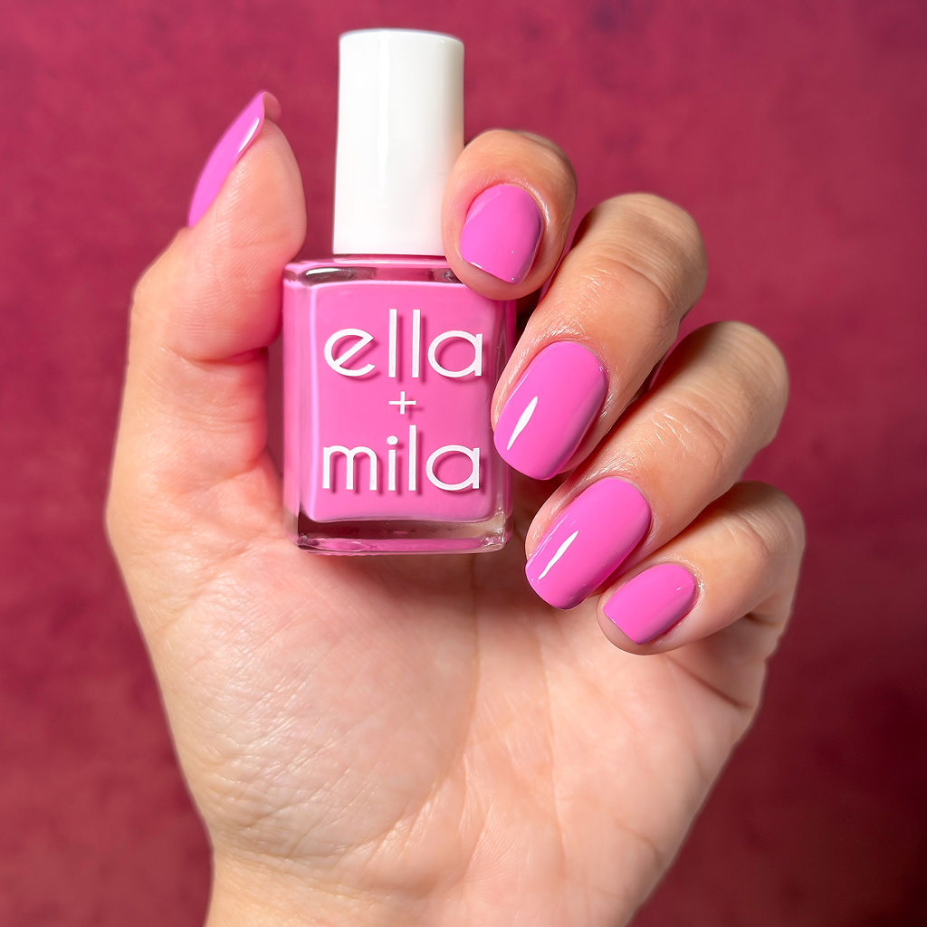 A hand with bright pink nails holds a matching bottle of Ella + Mila nail polish.