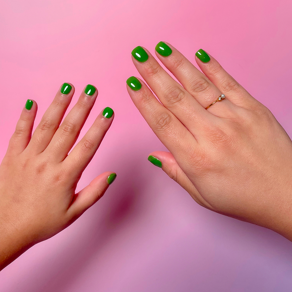 Two hands with bright green manicured nails, one with a ring, against a pink and purple background.